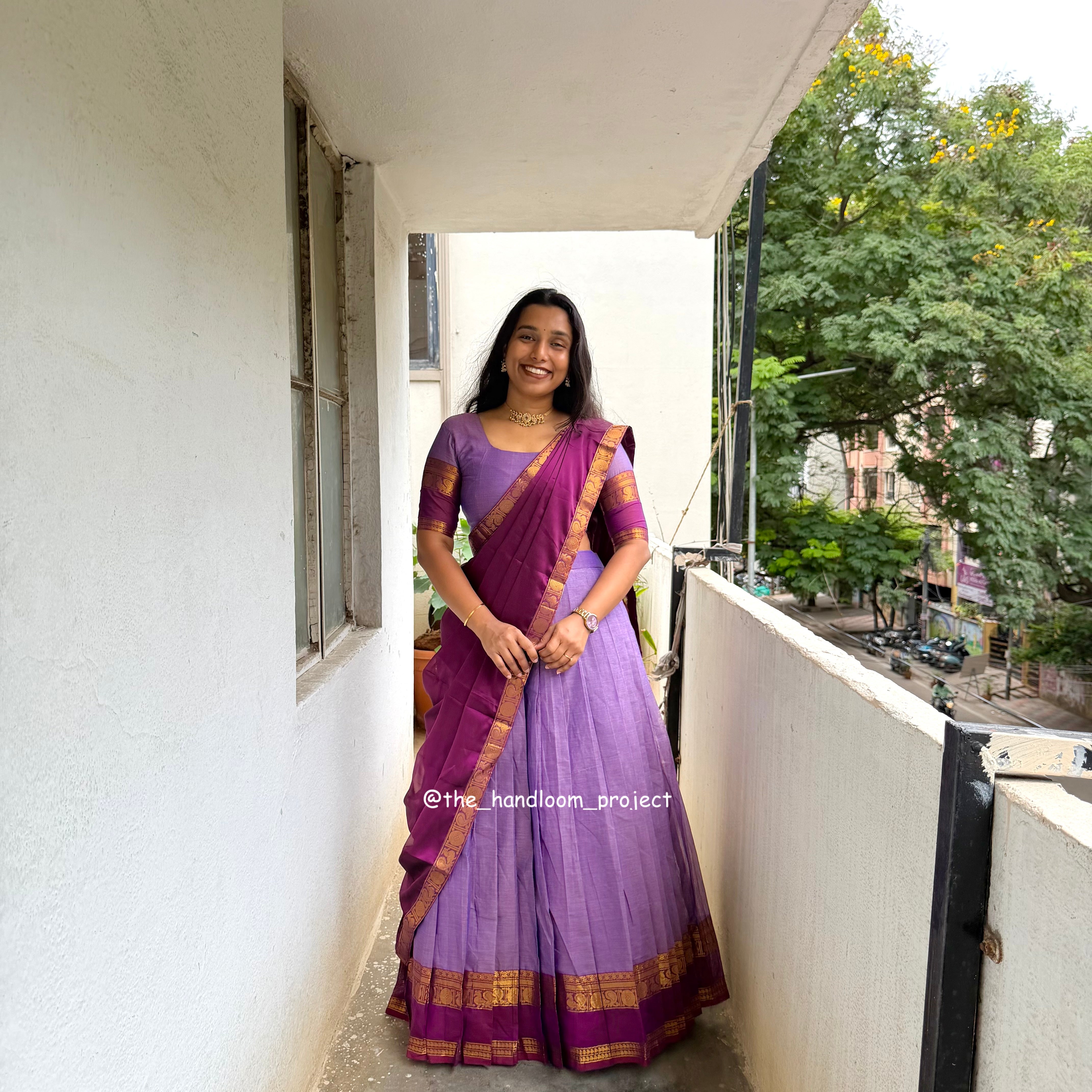 A woman stands on a white-walled balcony wearing a purple traditional sari with gold zari border detailing.