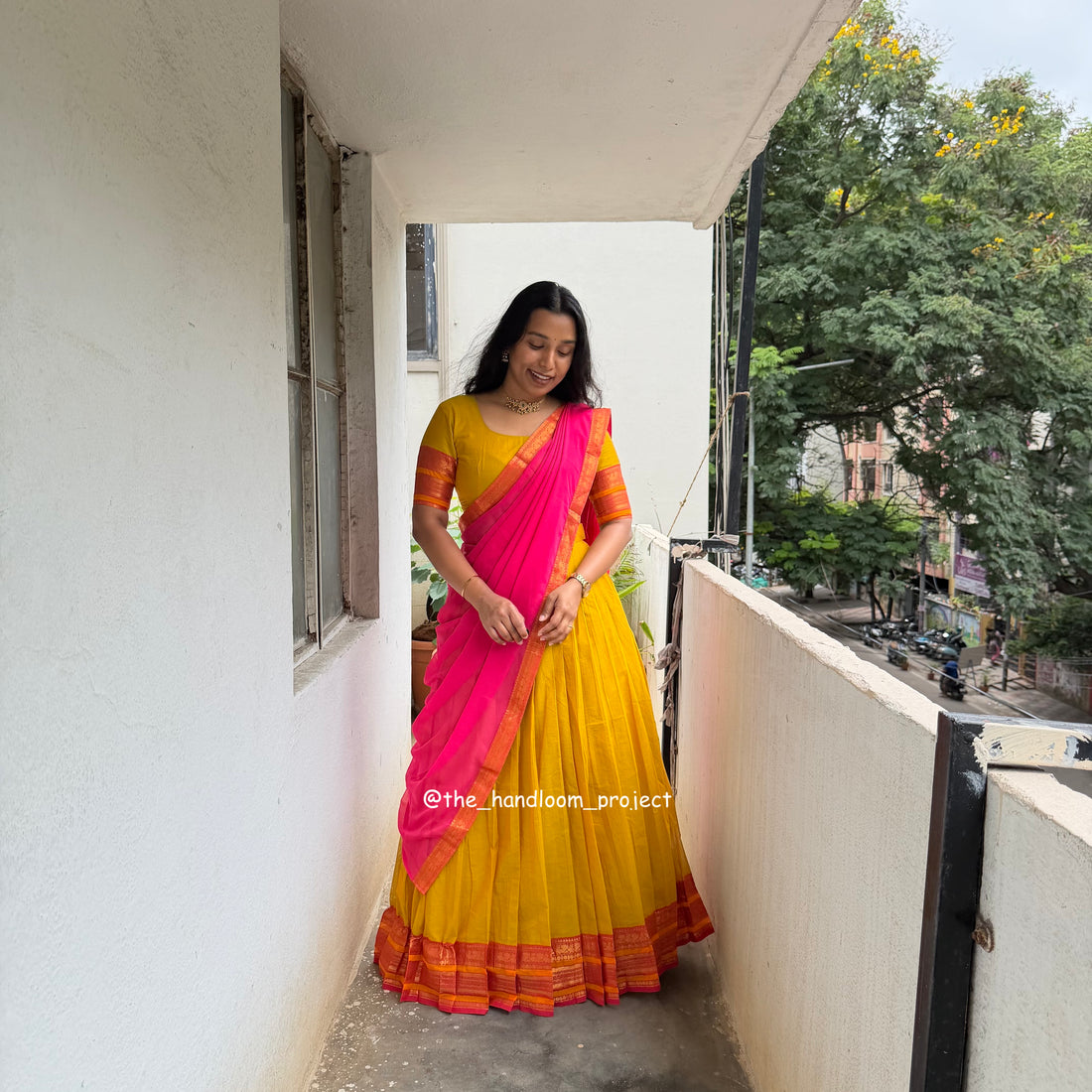 Woman wearing a traditional Indian outfit with a yellow skirt, orange blouse, and bright pink dupatta, standing on a balcony.